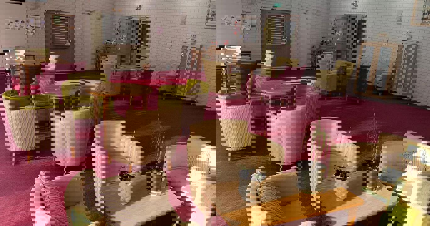 A sun-lit recreation room with a red carpet, white walls and a number of comfortable chairs surrounding coffee tables